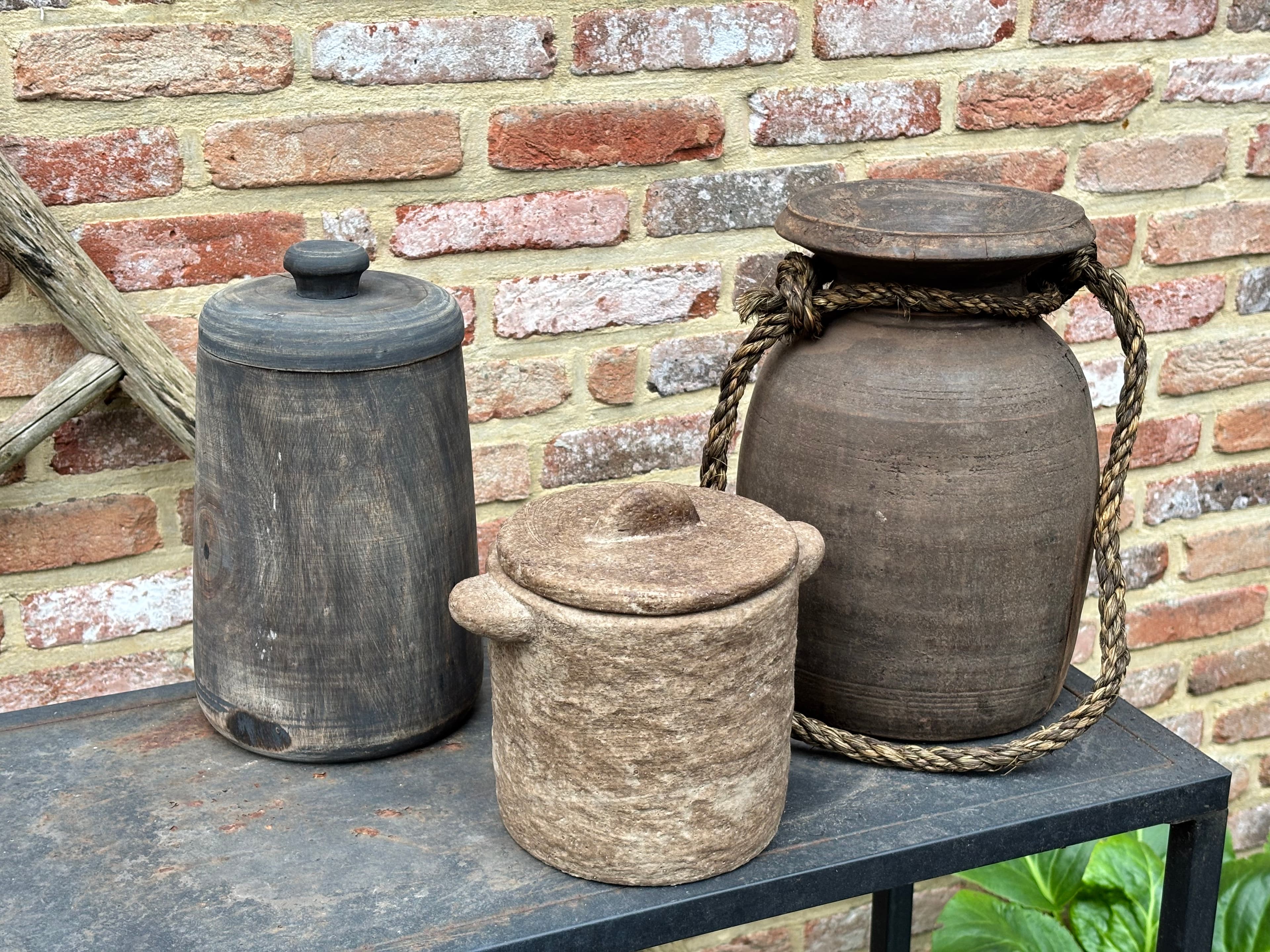 Three rustic wooden and stone vessels on a metal table against a weathered brick wall.