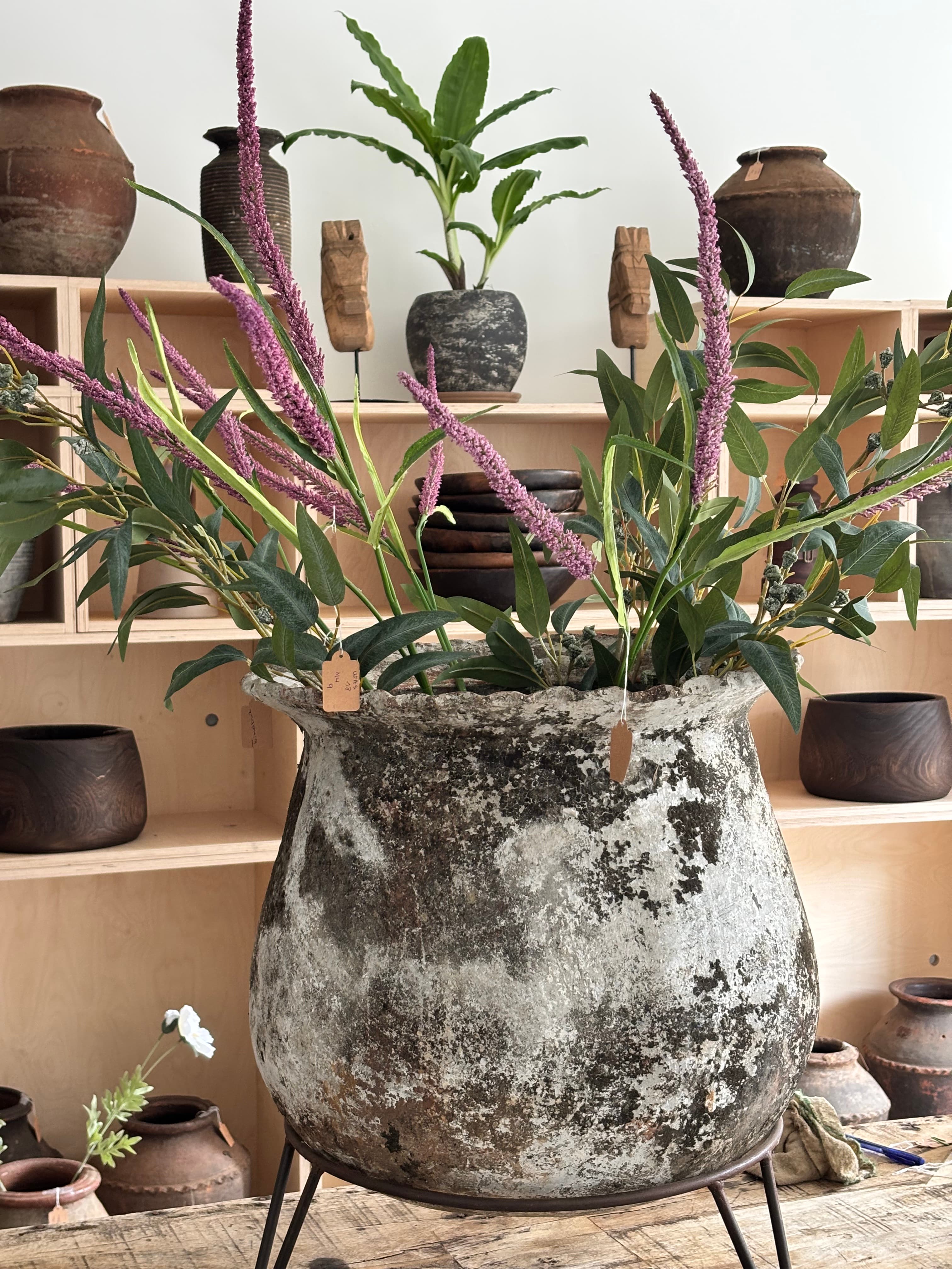 Large weathered rustic pot with purple flowers and green leaves against wooden display shelves.