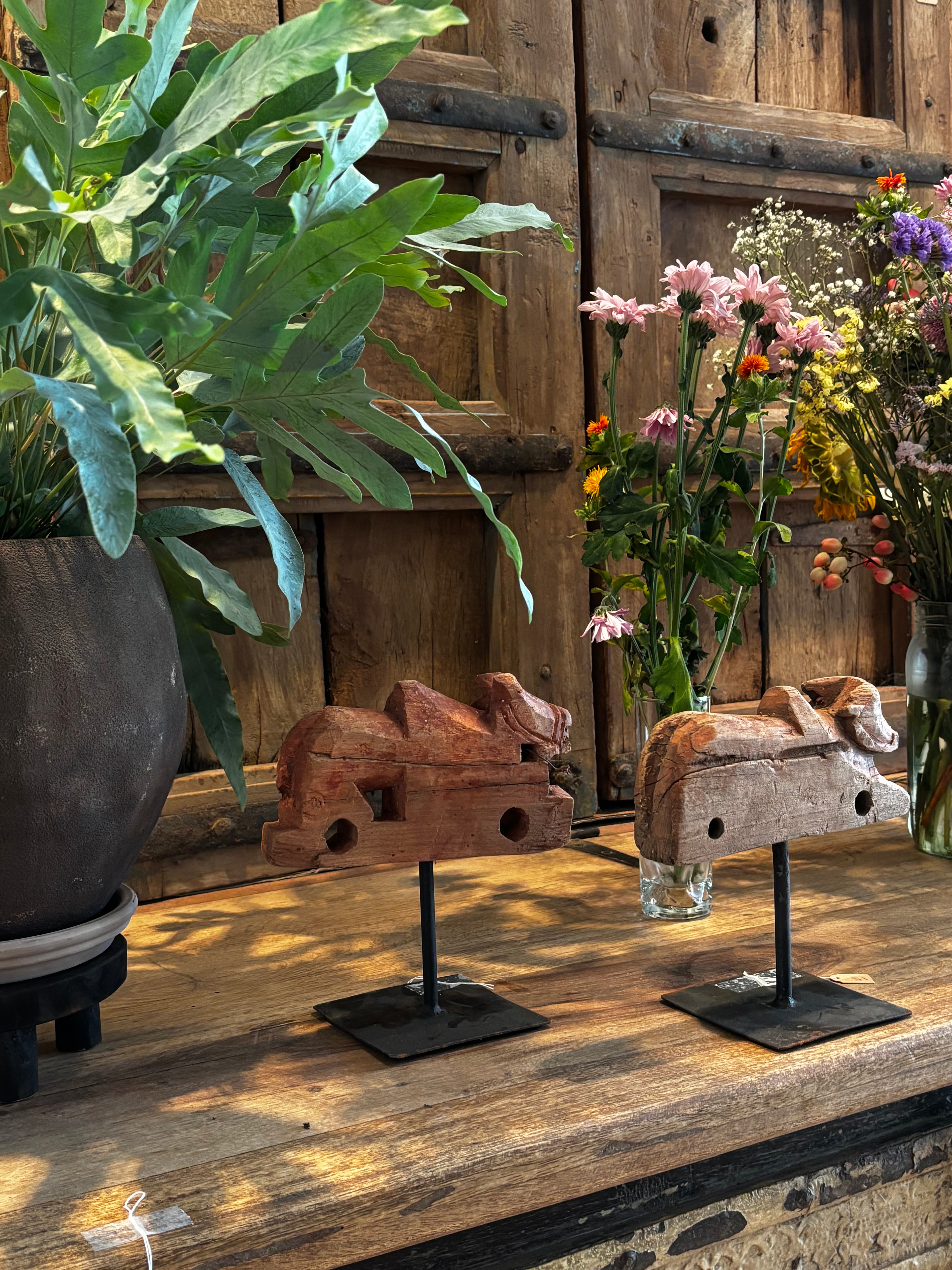 Two rustic wooden animal carvings on stands between a potted plant and colorful flowers.