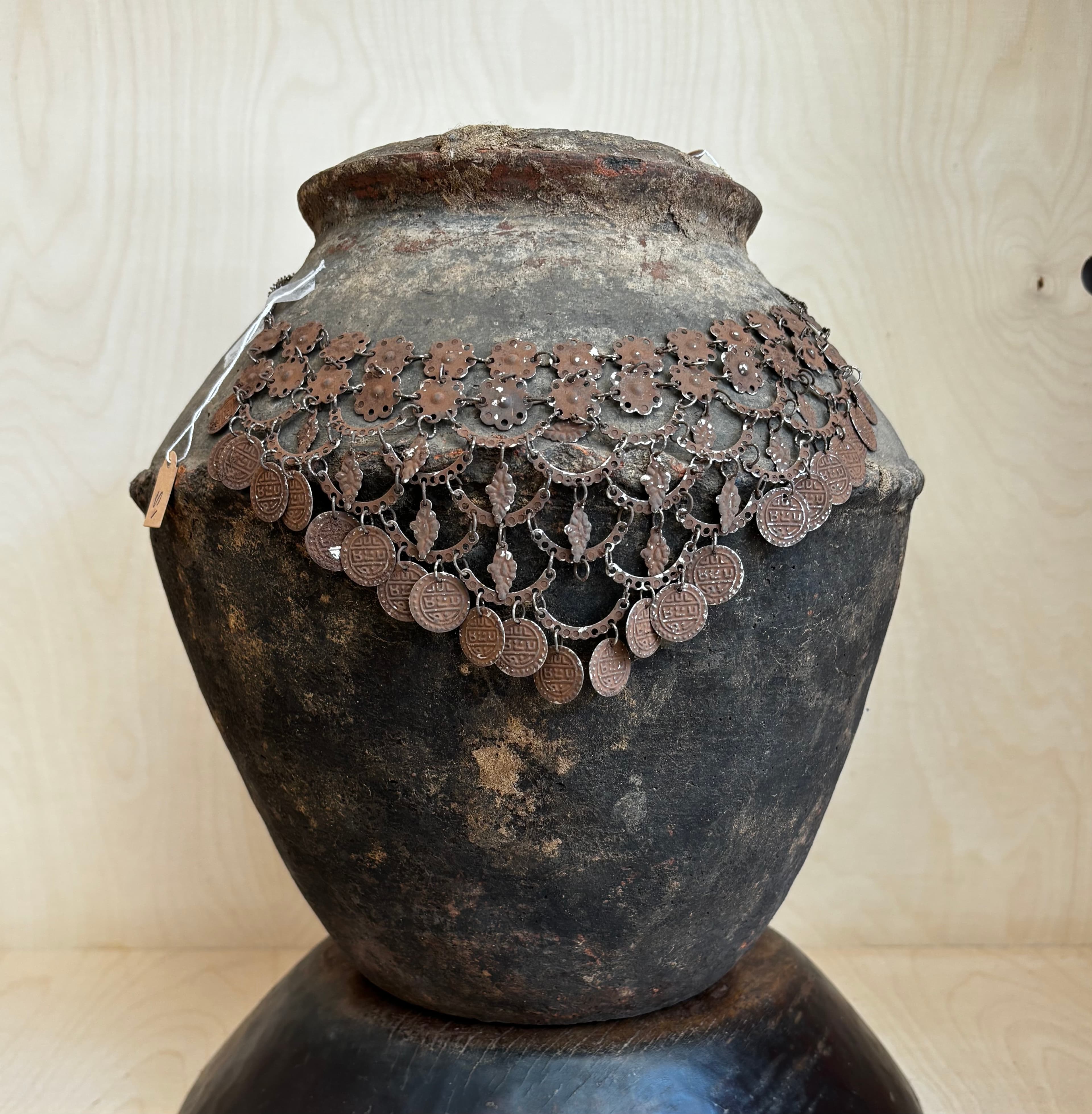 Ancient dark ceramic jar draped with an ornate metal coin necklace against light wood.
