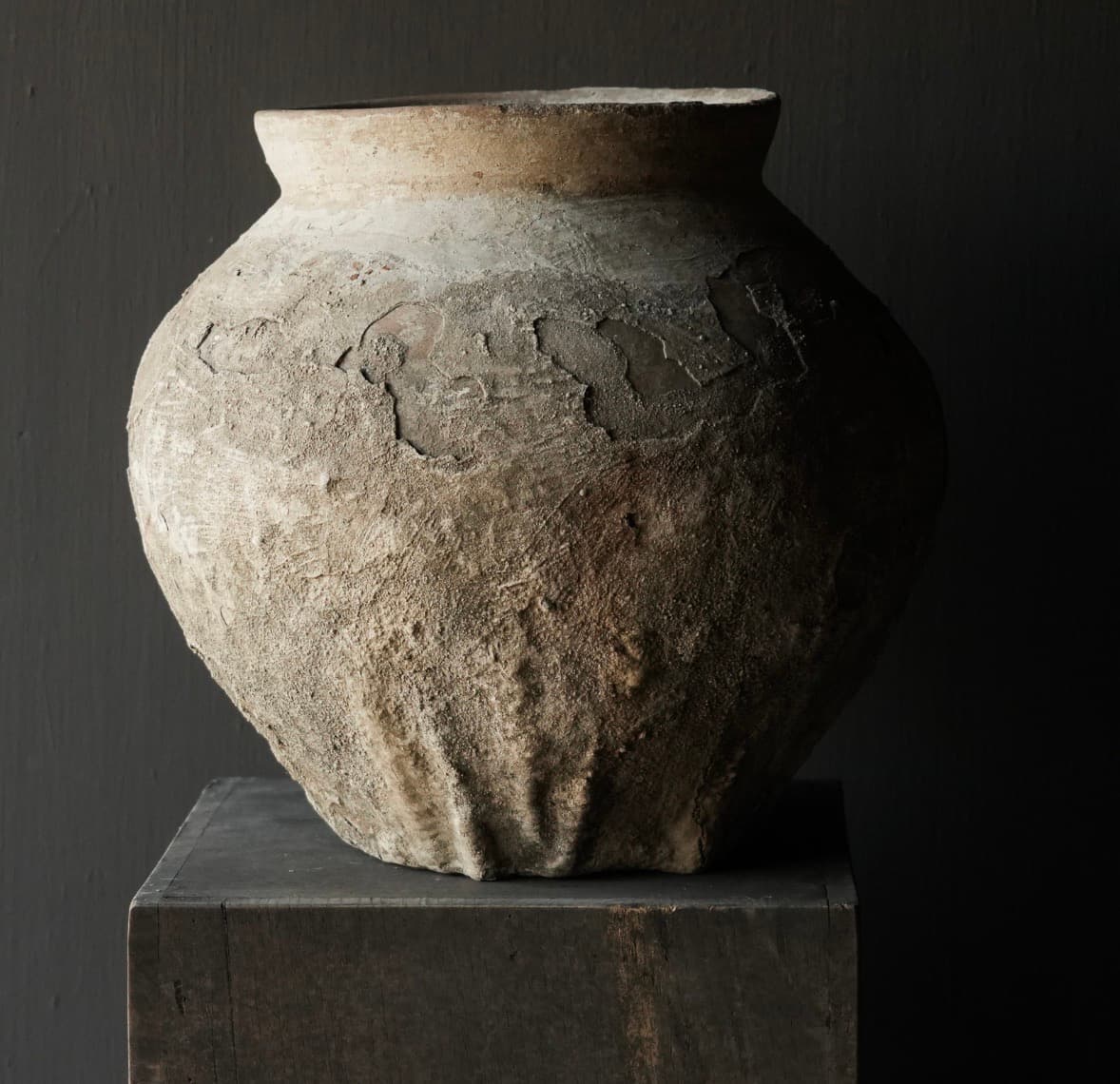 Ancient bulbous clay pot with a heavily weathered, peeling surface on a dark wooden pedestal.