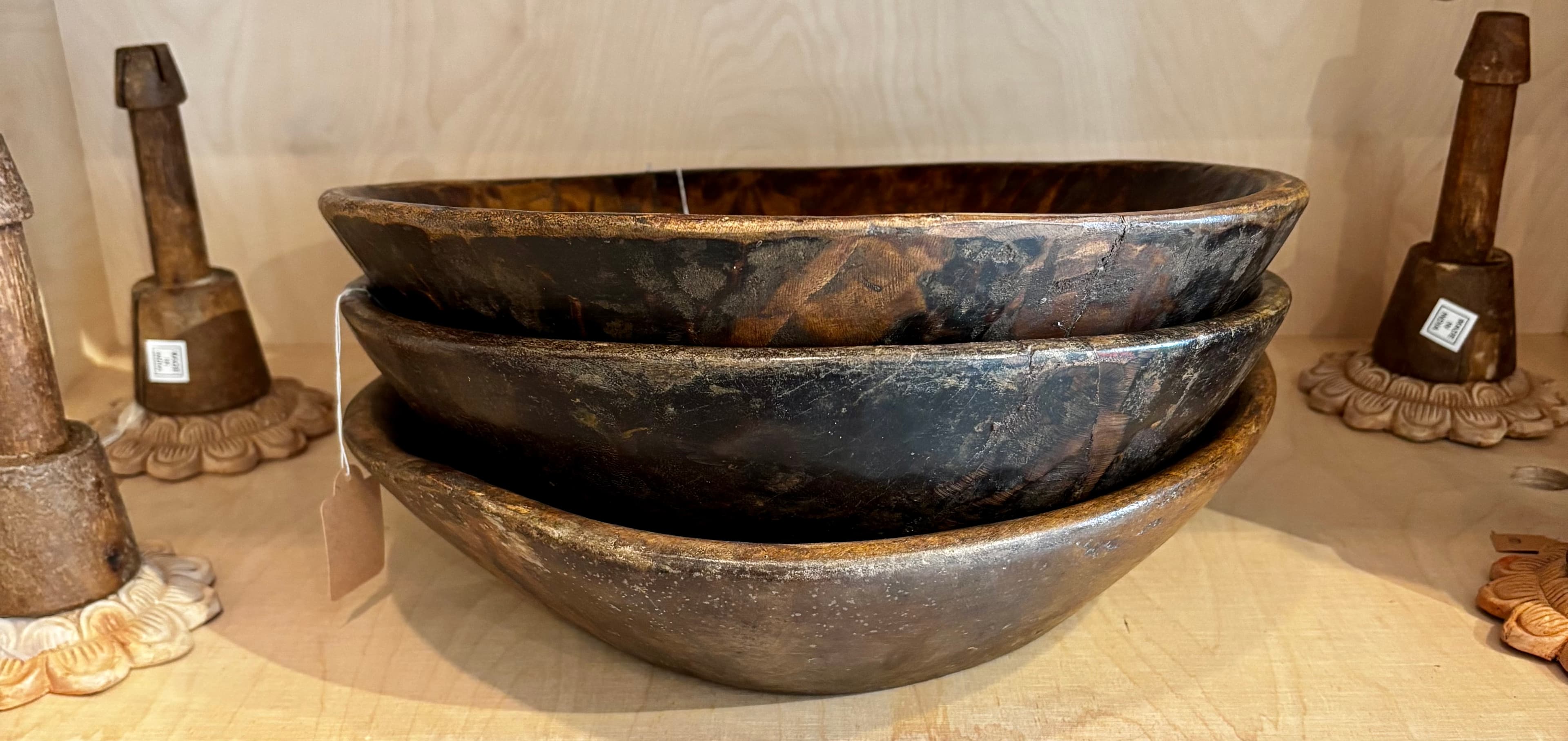 Three rustic dark wood bowls stacked on a light shelf flanked by carved wooden stands.