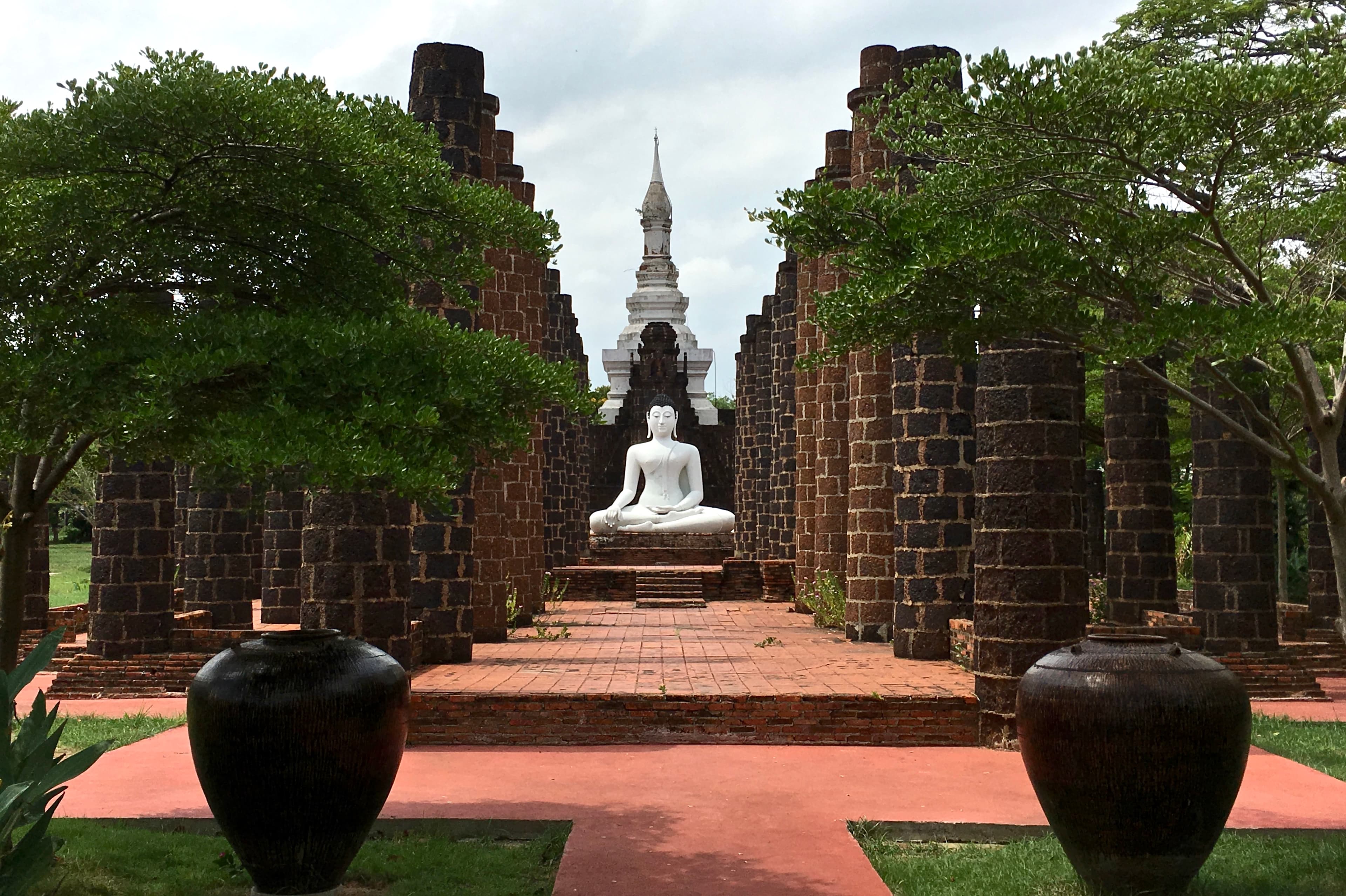 White seated Buddha statue centered between ancient brick pillars with a white stupa behind.