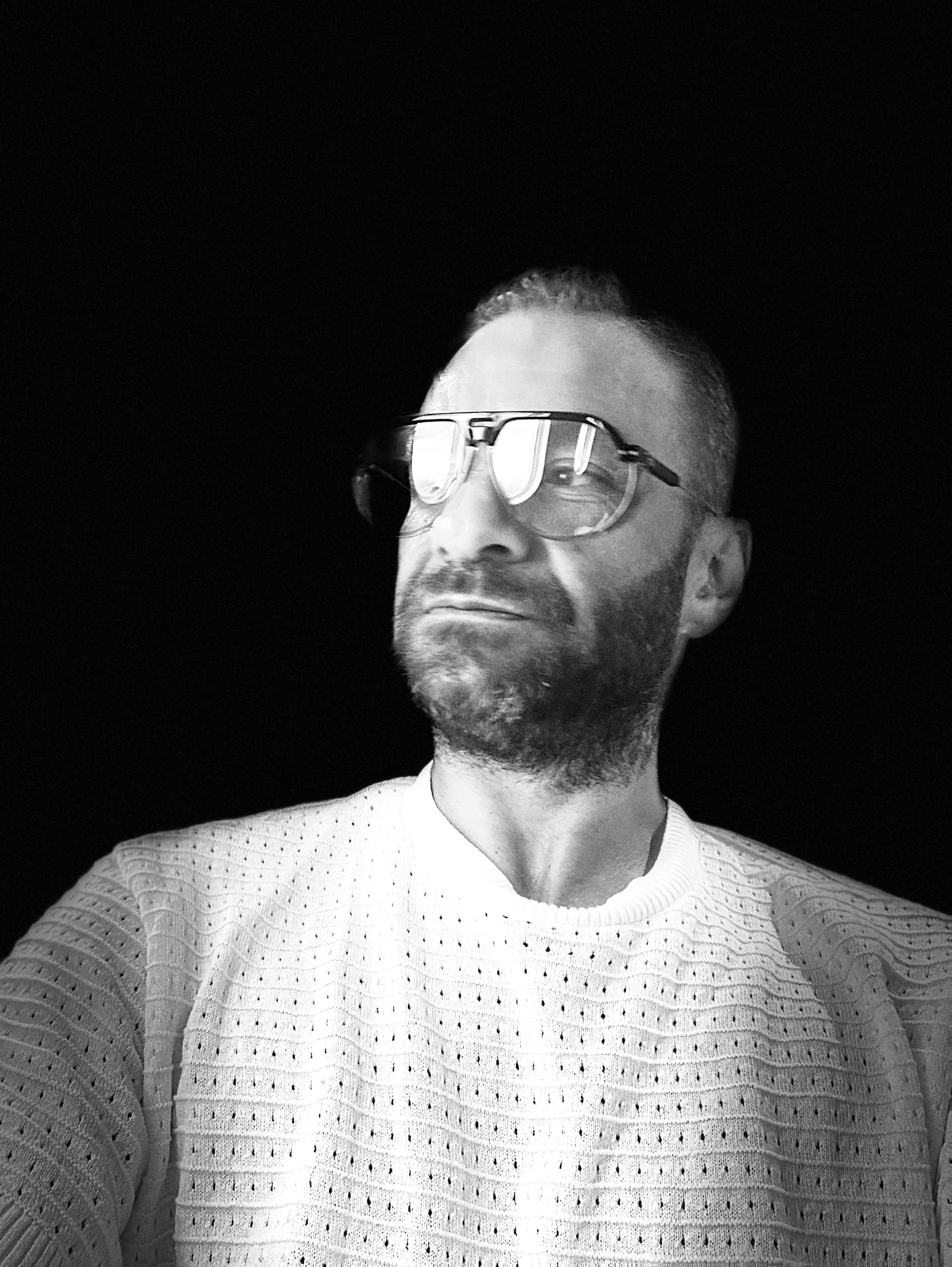 Bearded man in large glasses and textured white sweater looking away against a black background.