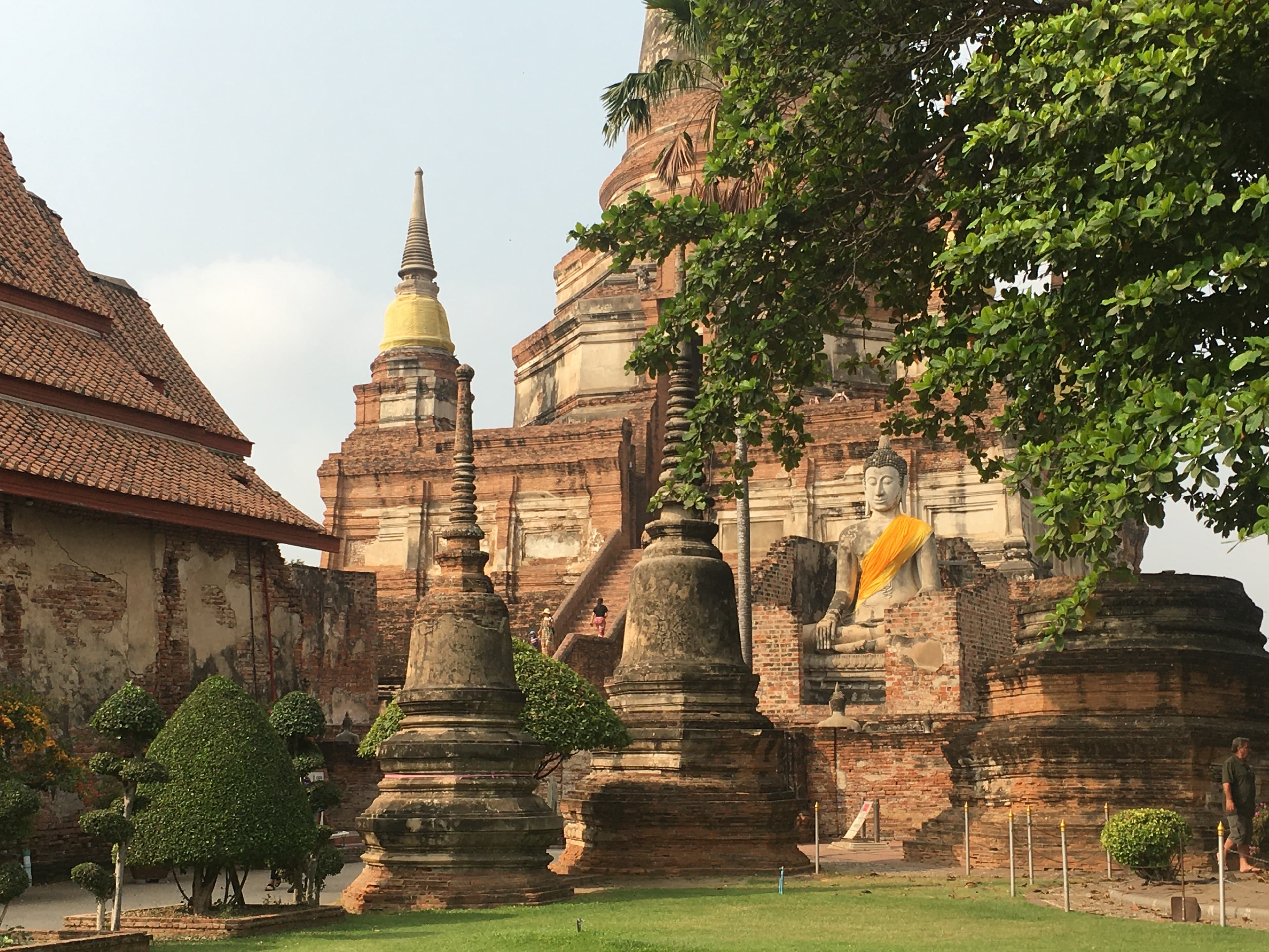 Large seated Buddha statue with yellow sash among ancient brick stupas at a Thai temple.